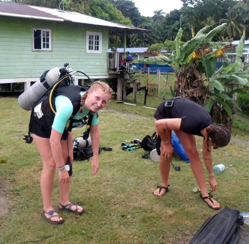 Two women are preparing for a scuba diving session. They are bending over, possibly adjusting their gear, on a grassy area. Both are wearing dark-colored tops and sandals. One has a scuba tank and gear on her back. In the background, there's a light green building with white trim and a few trees.